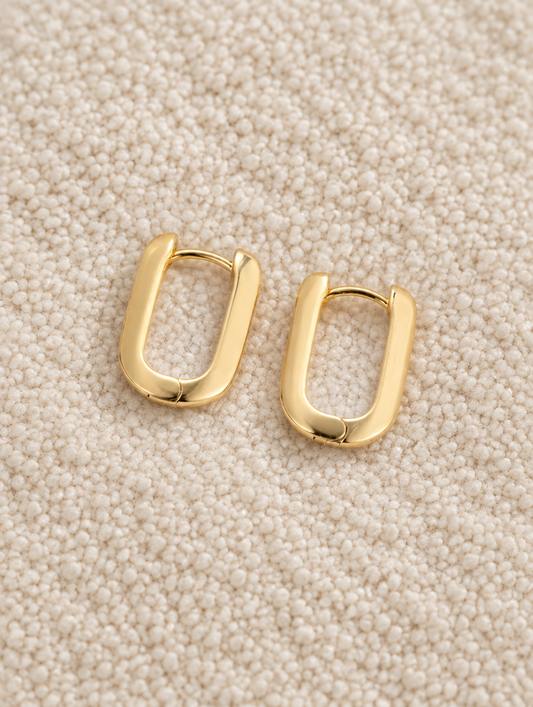 Gold-plated rounded rectangle huggie earrings lying flat on a soft styled surface