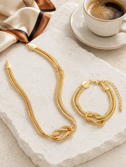 Gold knot necklace and bracelet on a stone slab with a coffee cup and silk scarf