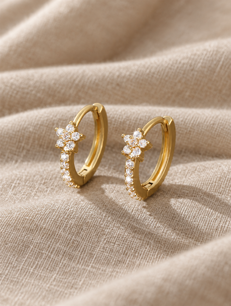 Gold huggie hoop earrings with cz flower cluster lying flat on a soft styled surface