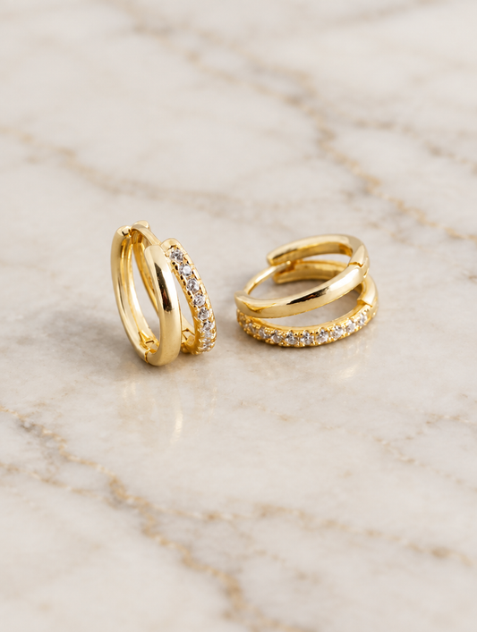Gold double-band huggie hoop earrings with cz stones lying flat on a soft styled surface
