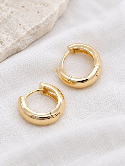Gold-plated chunky huggie hoop earrings lying flat on a soft styled surface