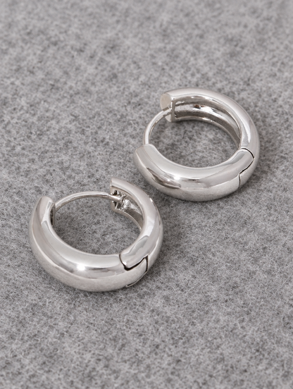 Silver-tone chunky huggie hoop earrings lying flat on a soft styled surface