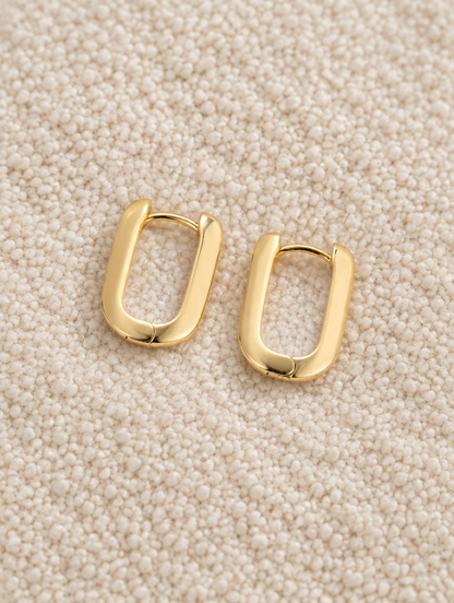 Gold-plated rounded rectangle huggie earrings lying flat on a soft styled surface