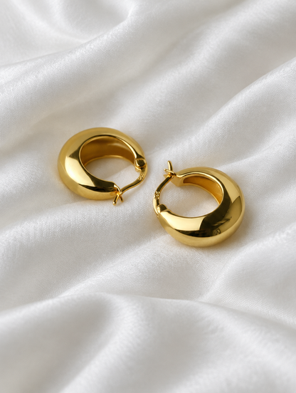 Gold-tone thick rounded hoop earrings lying flat on a soft styled surface