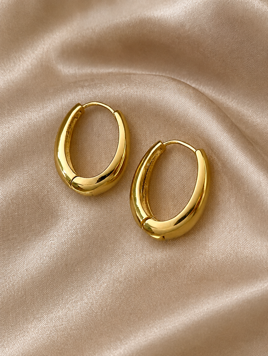 Slim gold-tone elongated oval hoop earrings lying flat on a soft styled surface