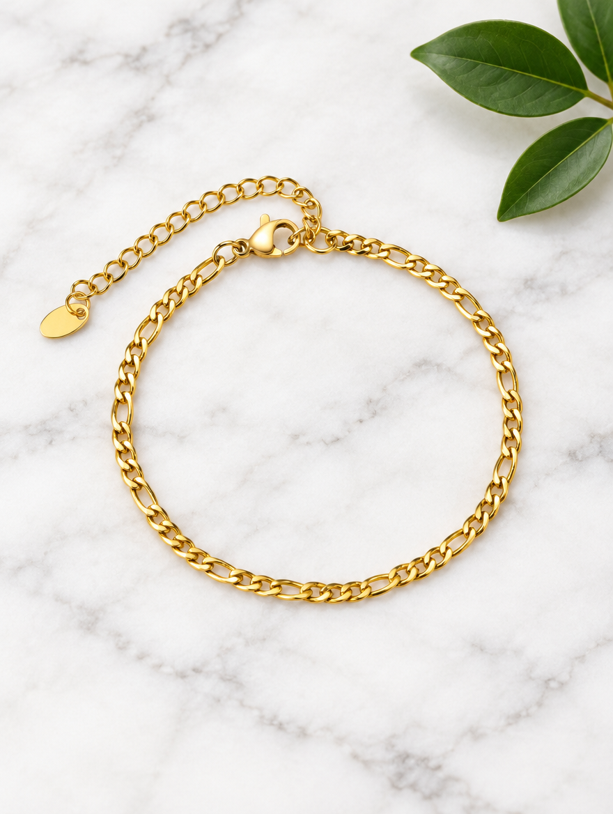 Gold figaro chain bracelet beside a green leaf on marble