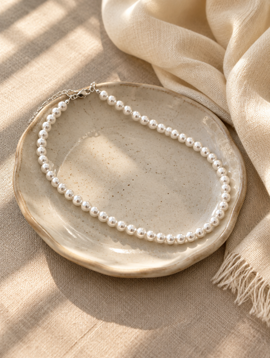 White pearl choker on a small ceramic dish beside a folded cream scarf