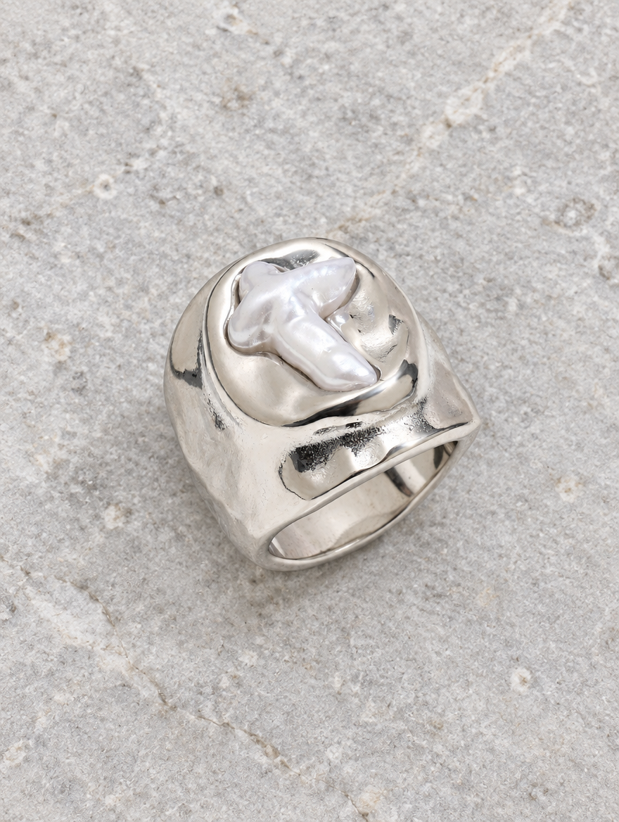 Silver chunky pearl cross ring resting on a soft textured surface