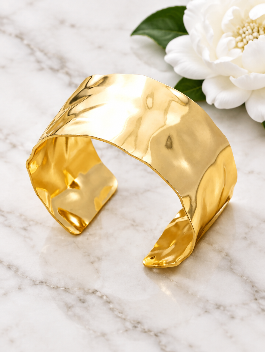 Wide gold cuff bracelet beside a white flower on marble