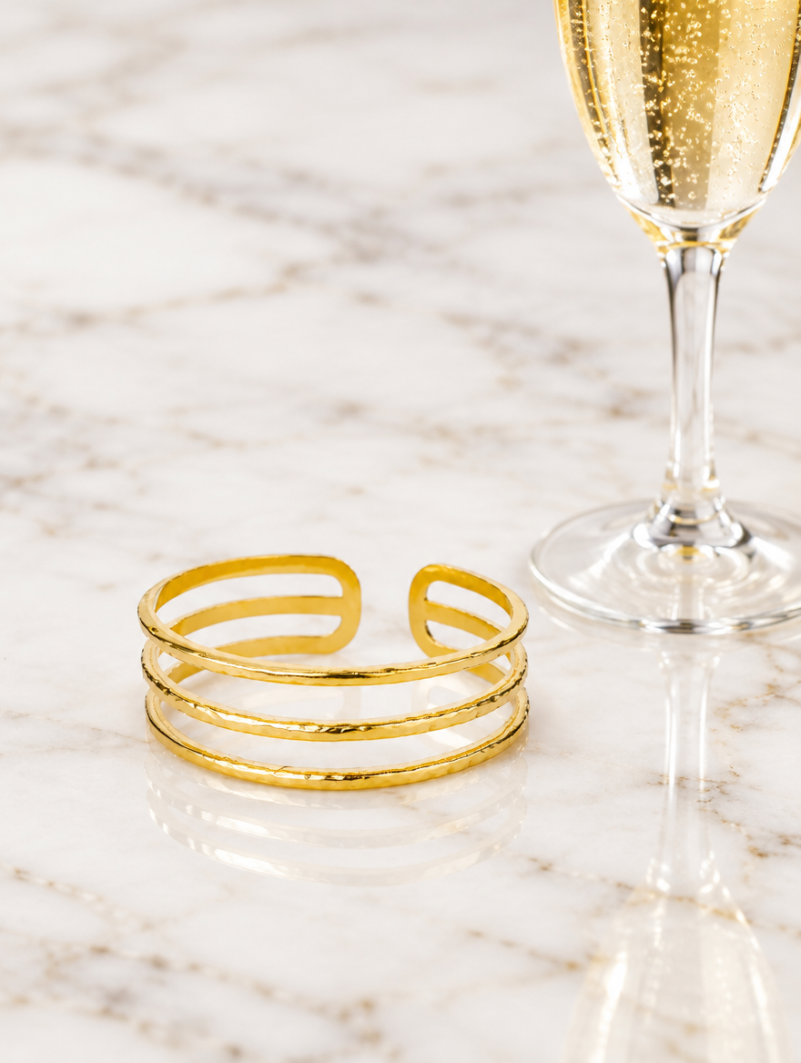 Gold triple line cuff beside a champagne flute on marble