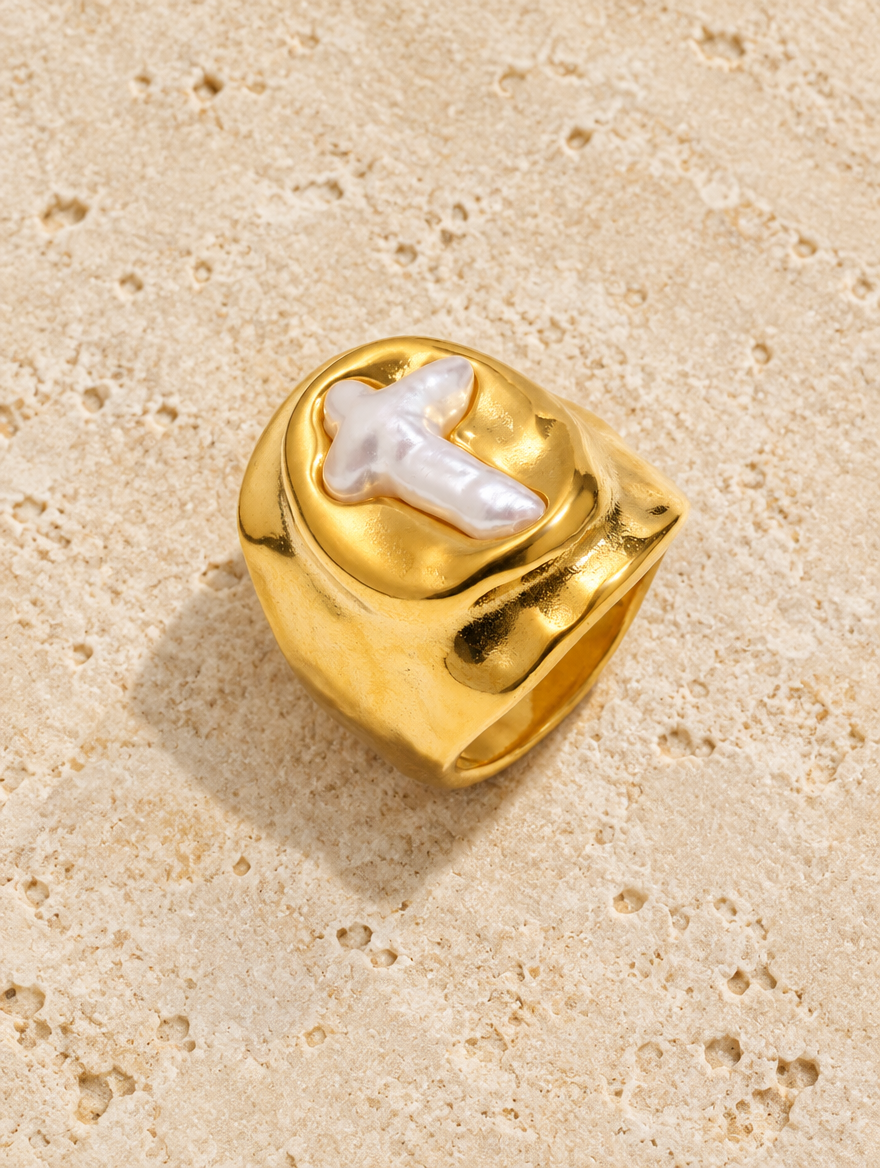 Gold chunky pearl cross ring resting on a soft textured surface