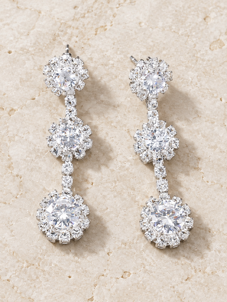 Silver three-flower crystal drop earrings lying flat on a soft styled surface