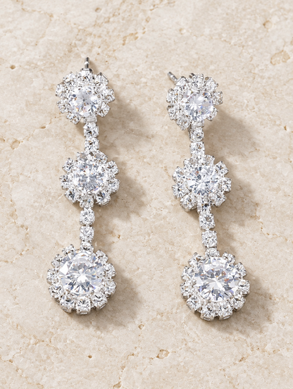 Silver three-flower crystal drop earrings lying flat on a soft styled surface
