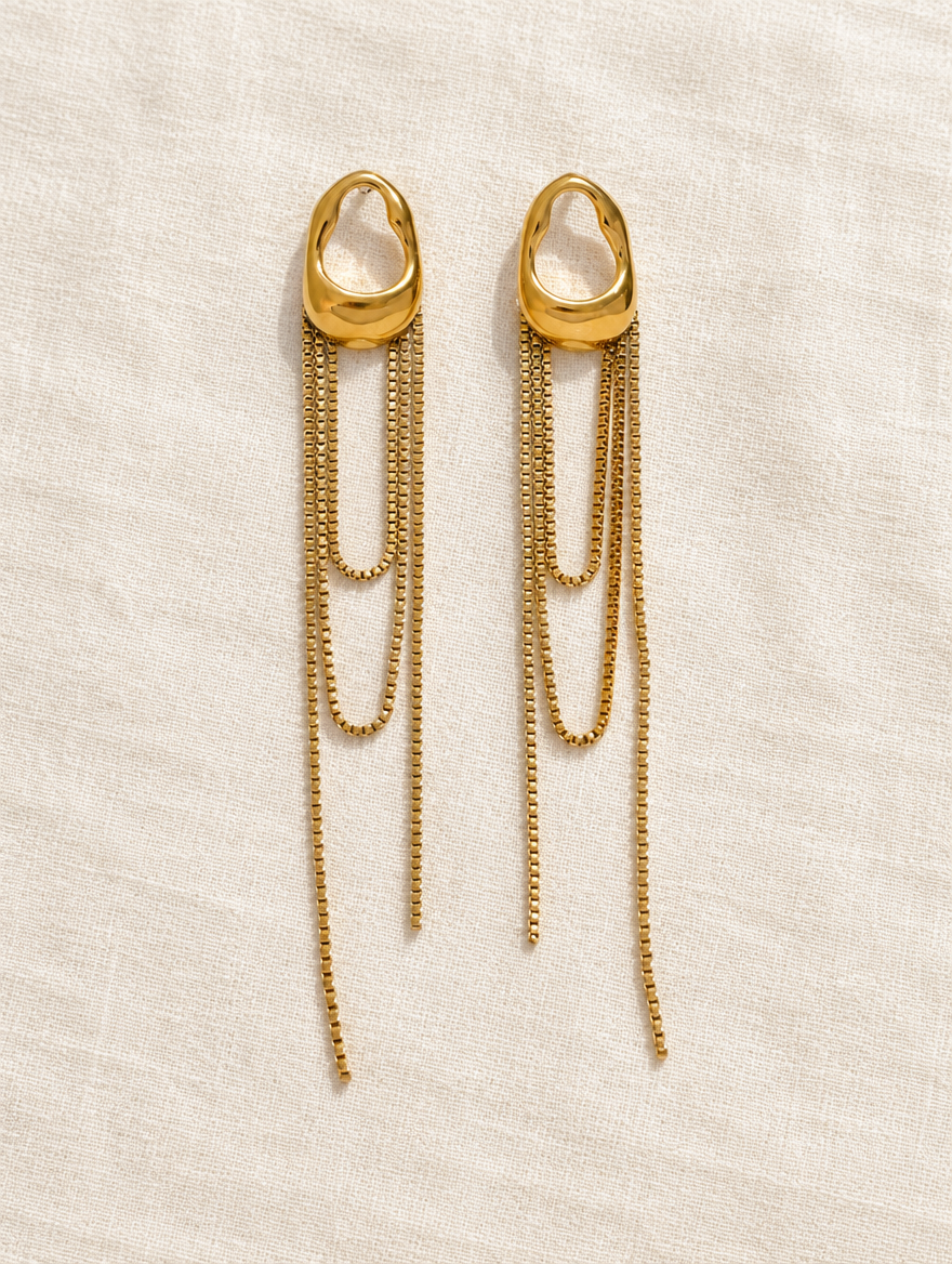 Gold chain tassel drop earrings with pearl-look bead lying flat on a soft styled surface