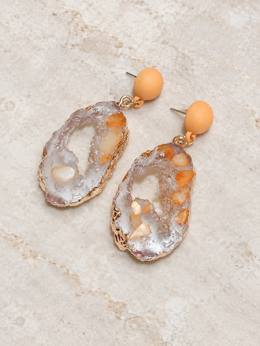 Orange stone-look drop earrings with gold-tone edge lying flat on a soft styled surface