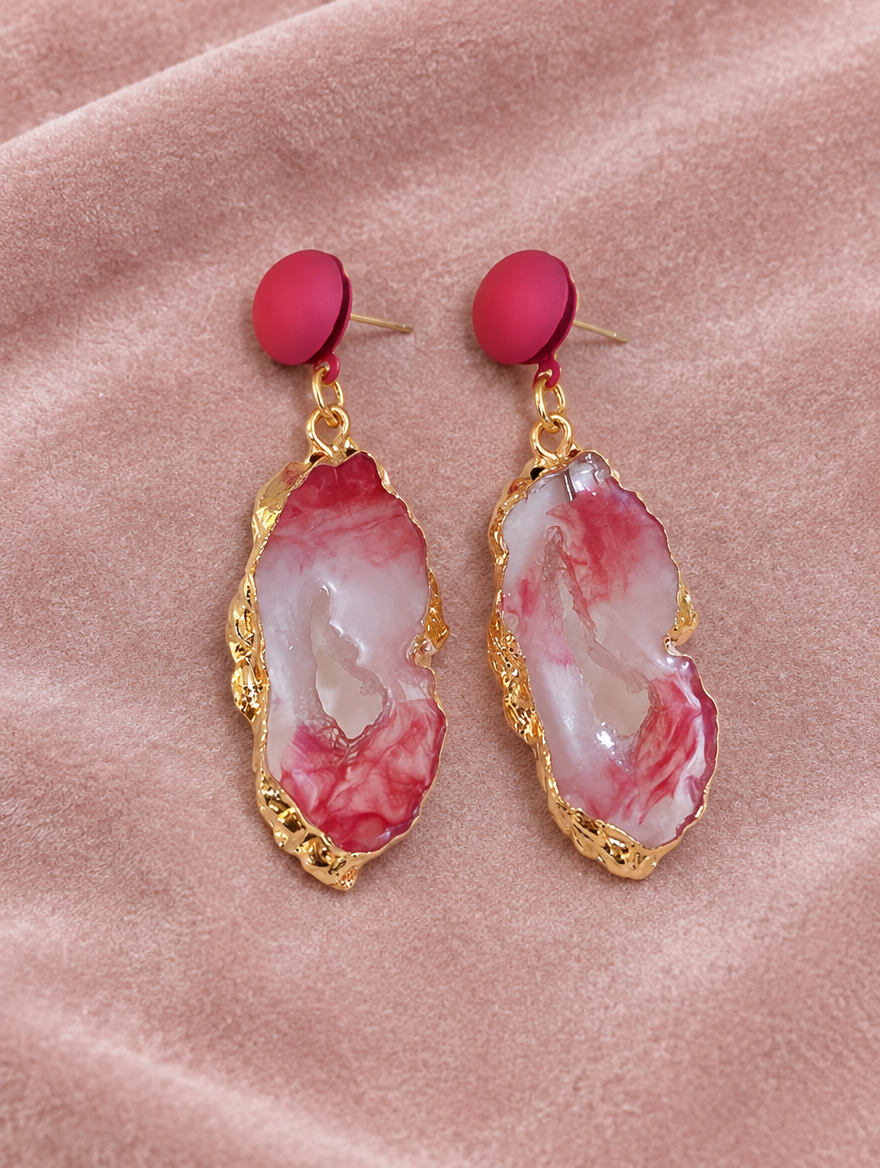 Pink stone-look drop earrings with gold-tone edge lying flat on a soft styled surface