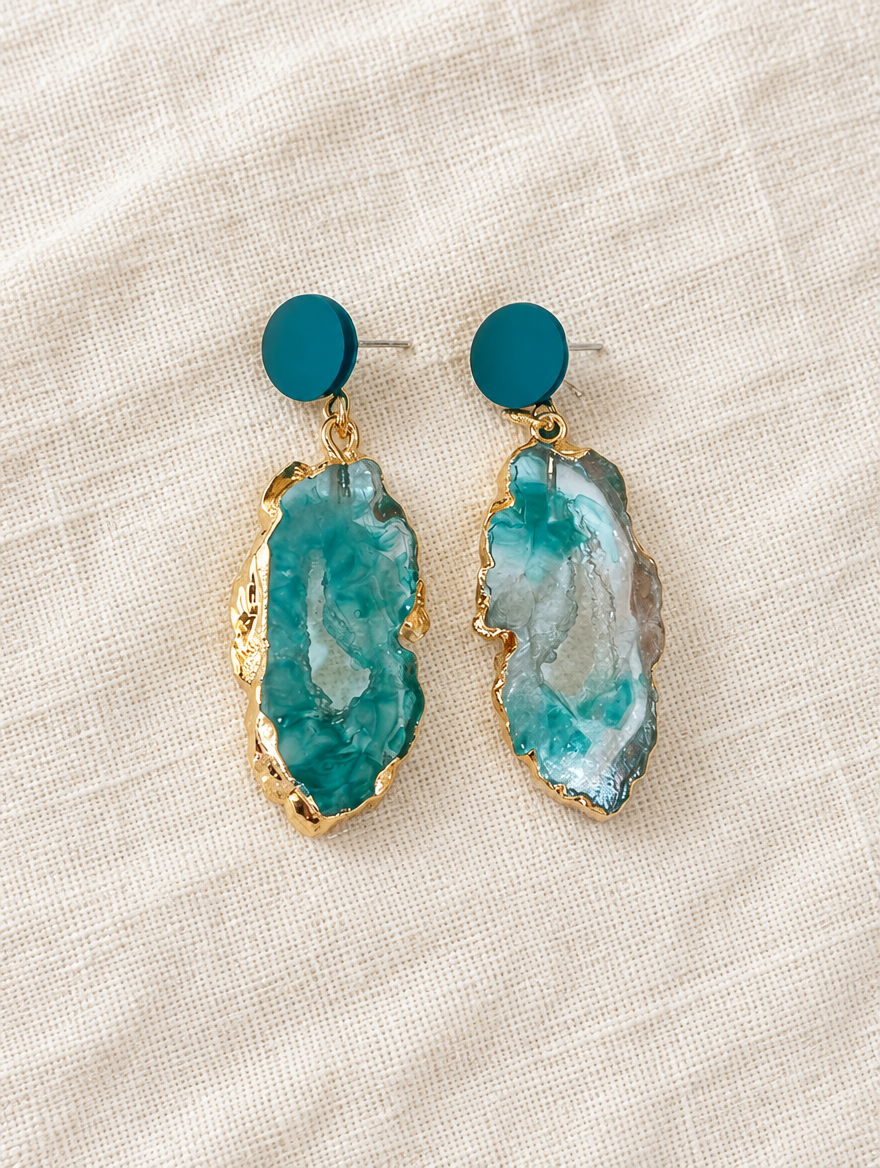 Teal stone-look drop earrings with gold-tone edge lying flat on a soft styled surface