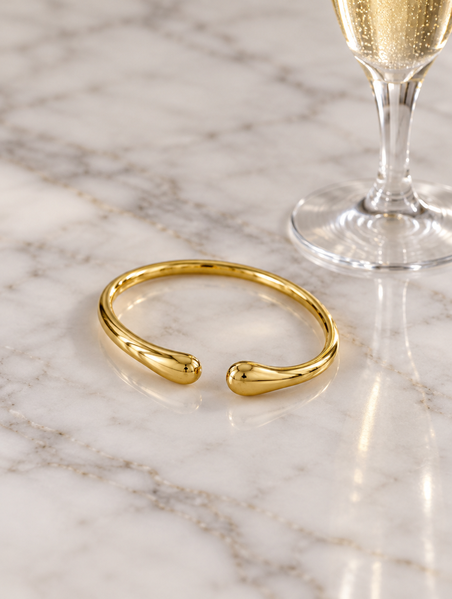 Slim gold-tone teardrop cuff beside a champagne flute on marble