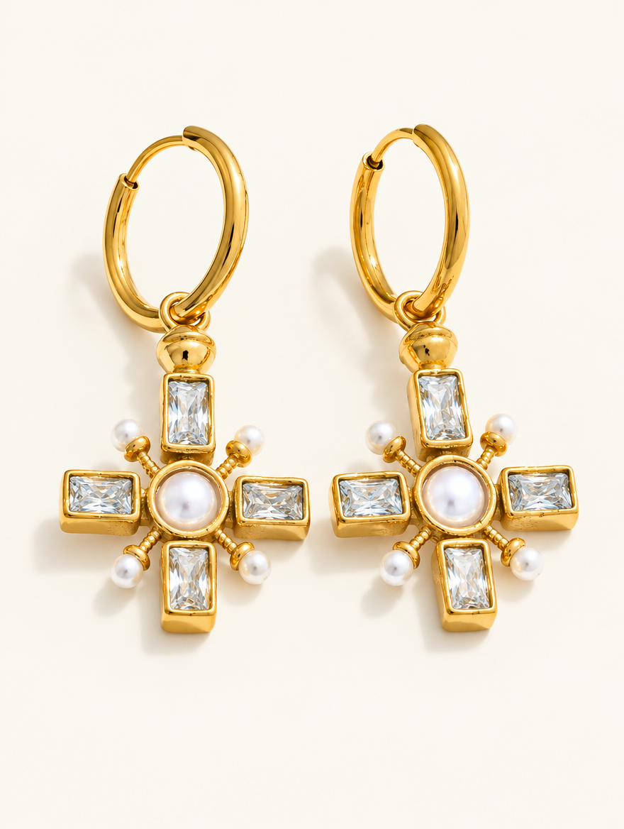 Pair of gold cross drop huggie earrings with pearl-look and CZ stones on a warm off-white background