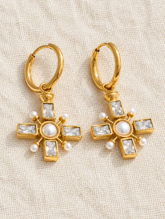 Gold cross drop huggie earrings with pearl-look and CZ stones lying flat on a soft styled surface