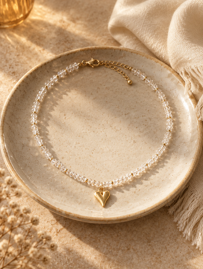 Clear bead choker with gold heart pendant on a ceramic dish beside a folded cream scarf
