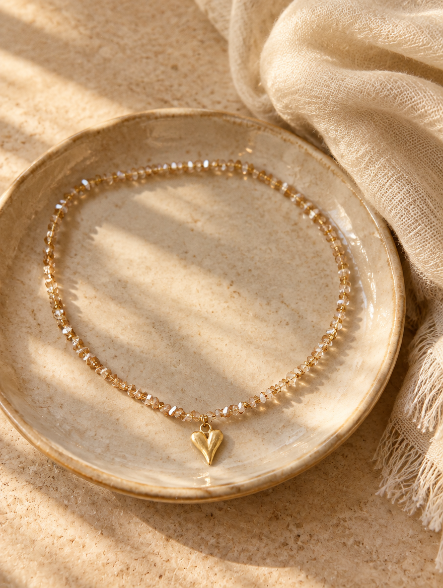Champagne bead choker with gold heart pendant on a ceramic dish beside a folded cream scarf