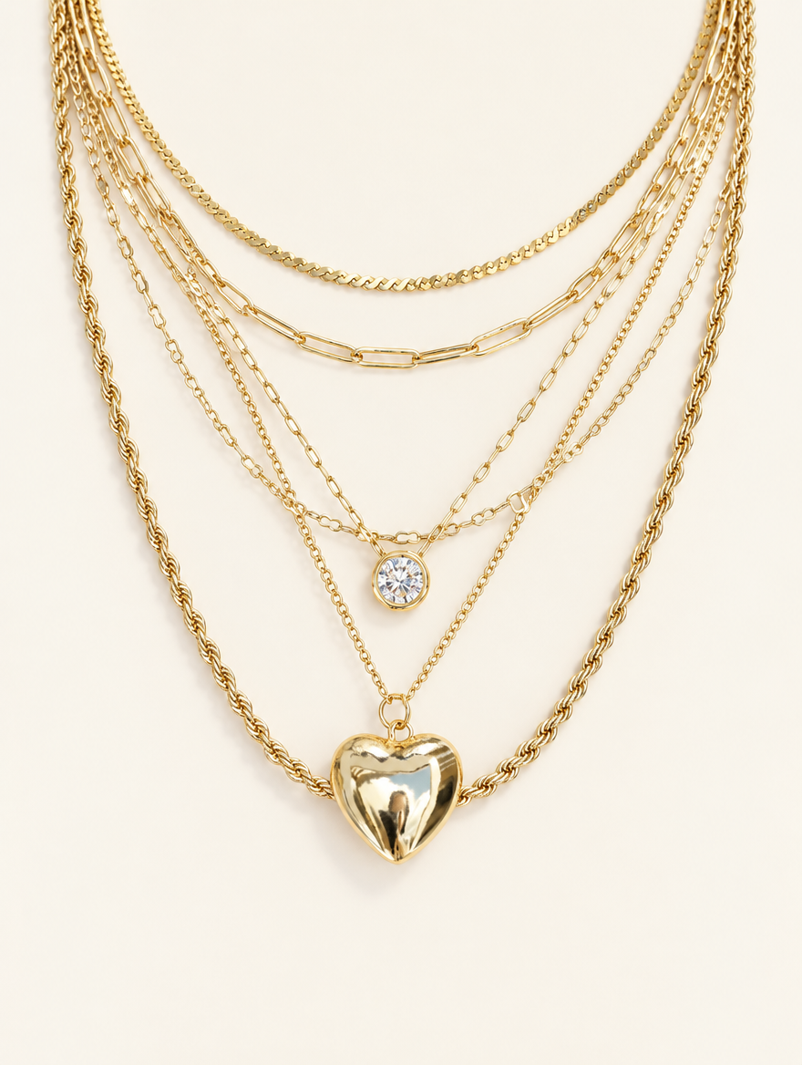 Gold layered necklace stack with a small crystal charm and a puffy gold heart pendant on a neutral background