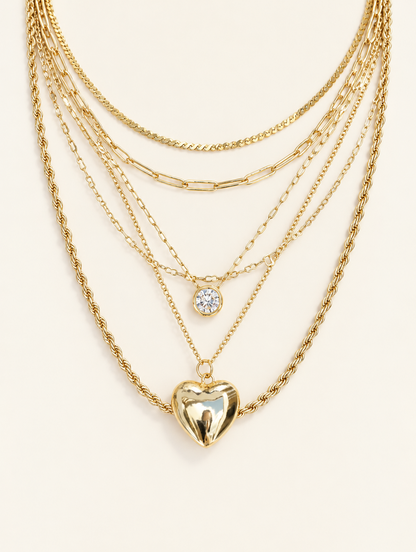 Gold layered necklace stack with a small crystal charm and a puffy gold heart pendant on a neutral background