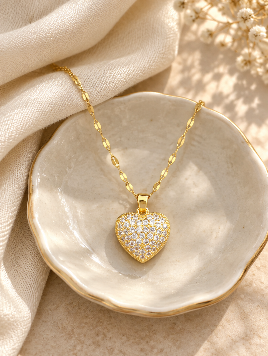 Gold pavé heart pendant necklace on a ceramic dish beside a folded cream scarf