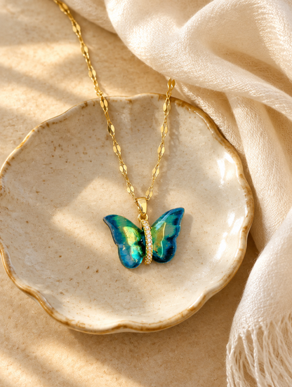 Gold enamel butterfly pendant necklace on a ceramic dish beside a folded cream scarf