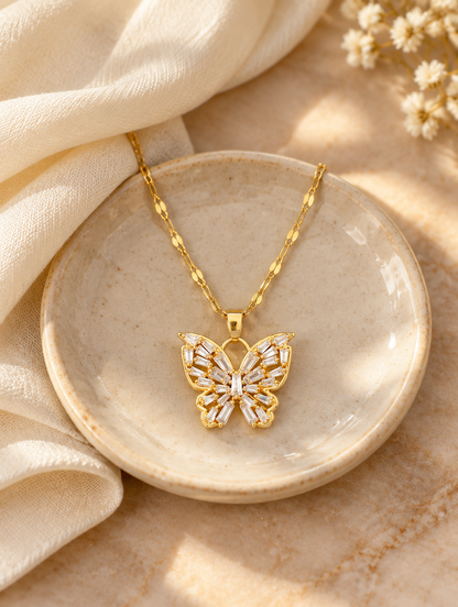 Gold crystal butterfly pendant necklace on a ceramic dish beside a folded cream scarf