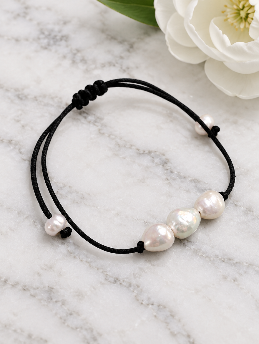 Black cord bracelet with baroque freshwater pearls beside a white flower on marble